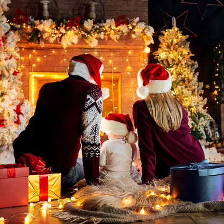 A cozy family moment by the fireplace — parents and child in Santa hats surrounded by twinkling lights, gifts, and a glowing Christmas tree — capturing the warmth and joy of the holiday season.